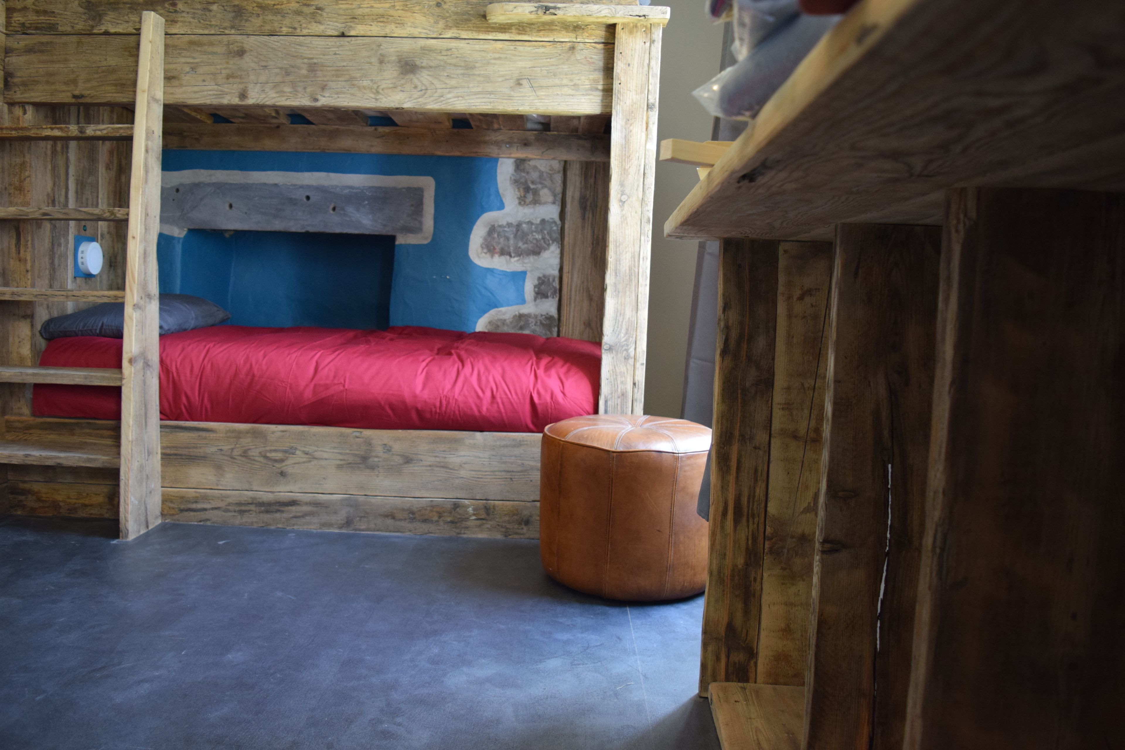 A rustic wooden bunk bed with a red mattress and a small round ottoman.