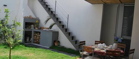 A modern outdoor patio with a dining table and chairs, a staircase, and a small garden area with plants and a tree.