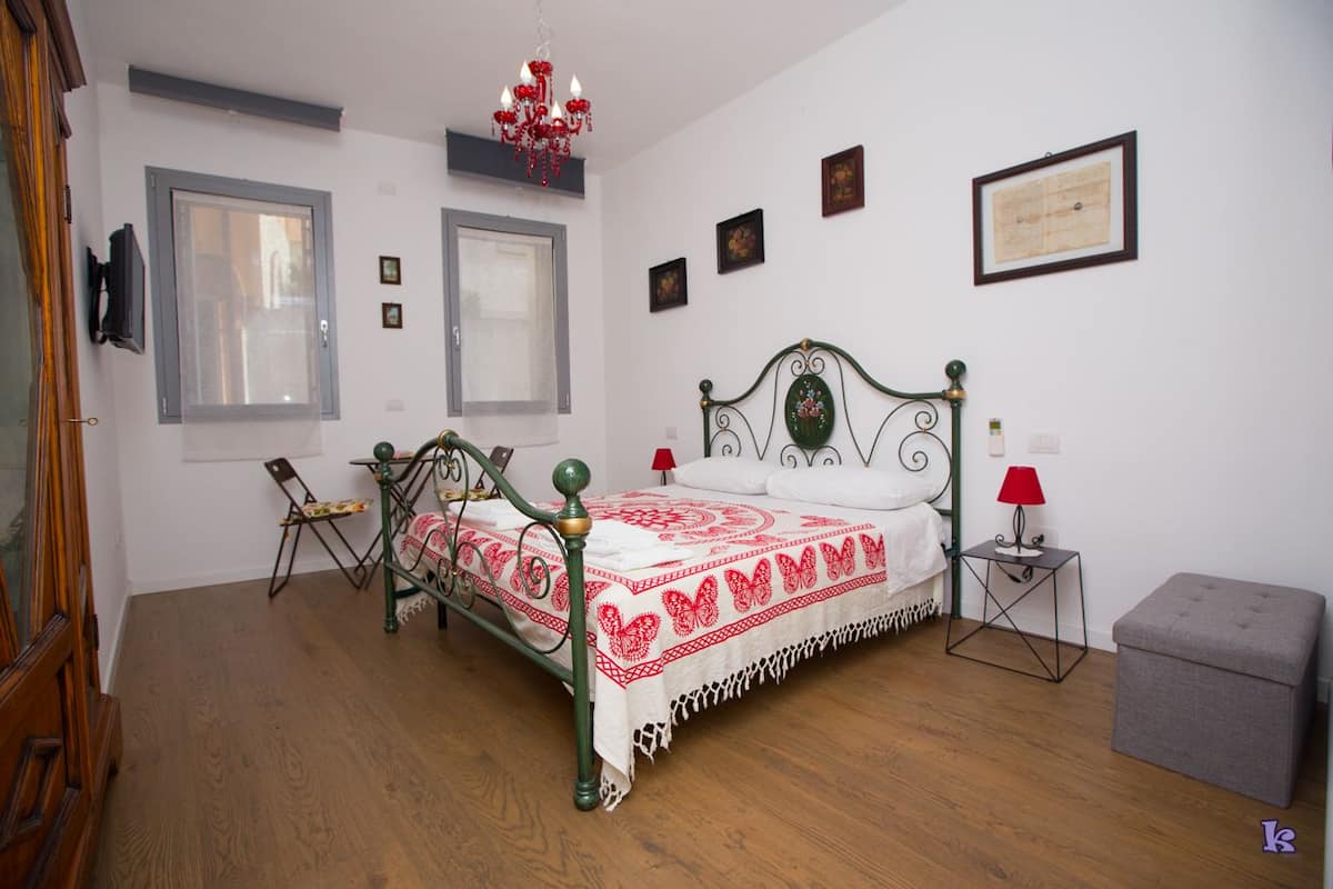 A bedroom with a wooden floor, a metal bed frame, a white bedspread with red patterns, two windows, a chandelier, a small table with a lamp, and a grey ottoman.