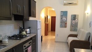 A compact kitchen with dark cabinets, a gas stove, a refrigerator, and a dining area with a brown sofa and a wooden table. - Maltese Rooms (Sliema)