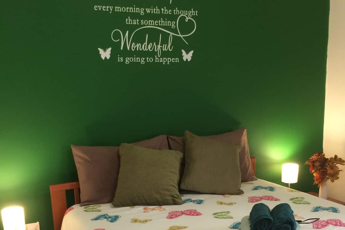 Ein Schlafzimmer mit grüner Wand, einem Bett mit weißer Decke mit bunten Schmetterlingsmustern, zwei Kissen, einem Paar zusammengerollter Handtücher, einer Nachttischlampe und einer Wandsticker mit einem motivierenden Zitat.