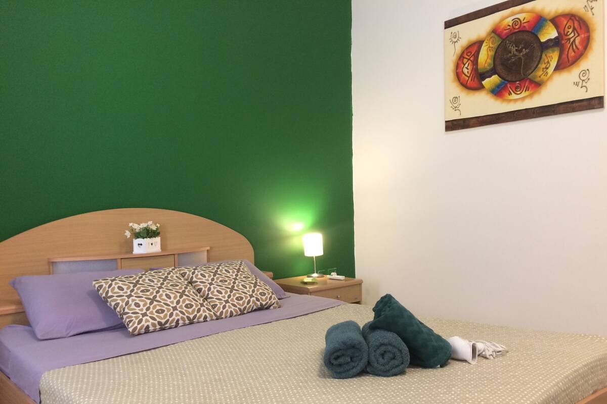 Ein ordentlich bezogenes Bett mit gemusterten Kissen, eine grüne Wand und ein Gemälde darüber.