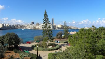 Un paisaje urbano costero con vista despejada al mar, un parque con bancos y varios edificios al fondo.