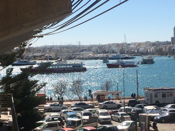 Un puerto deportivo con barcos amarrados, un estacionamiento con coches y un paisaje urbano al fondo.