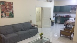 A living room with a grey sofa, a glass coffee table, and a small plant. There is a kitchen area with a table and chairs visible in the background. - Maltese Rooms (Sliema)