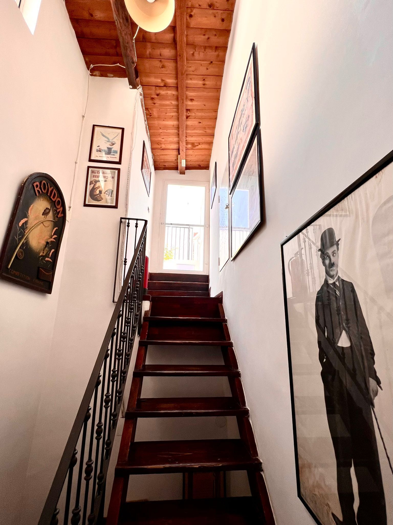 A wooden staircase with a metal railing leading to a bright room with framed pictures on the walls.