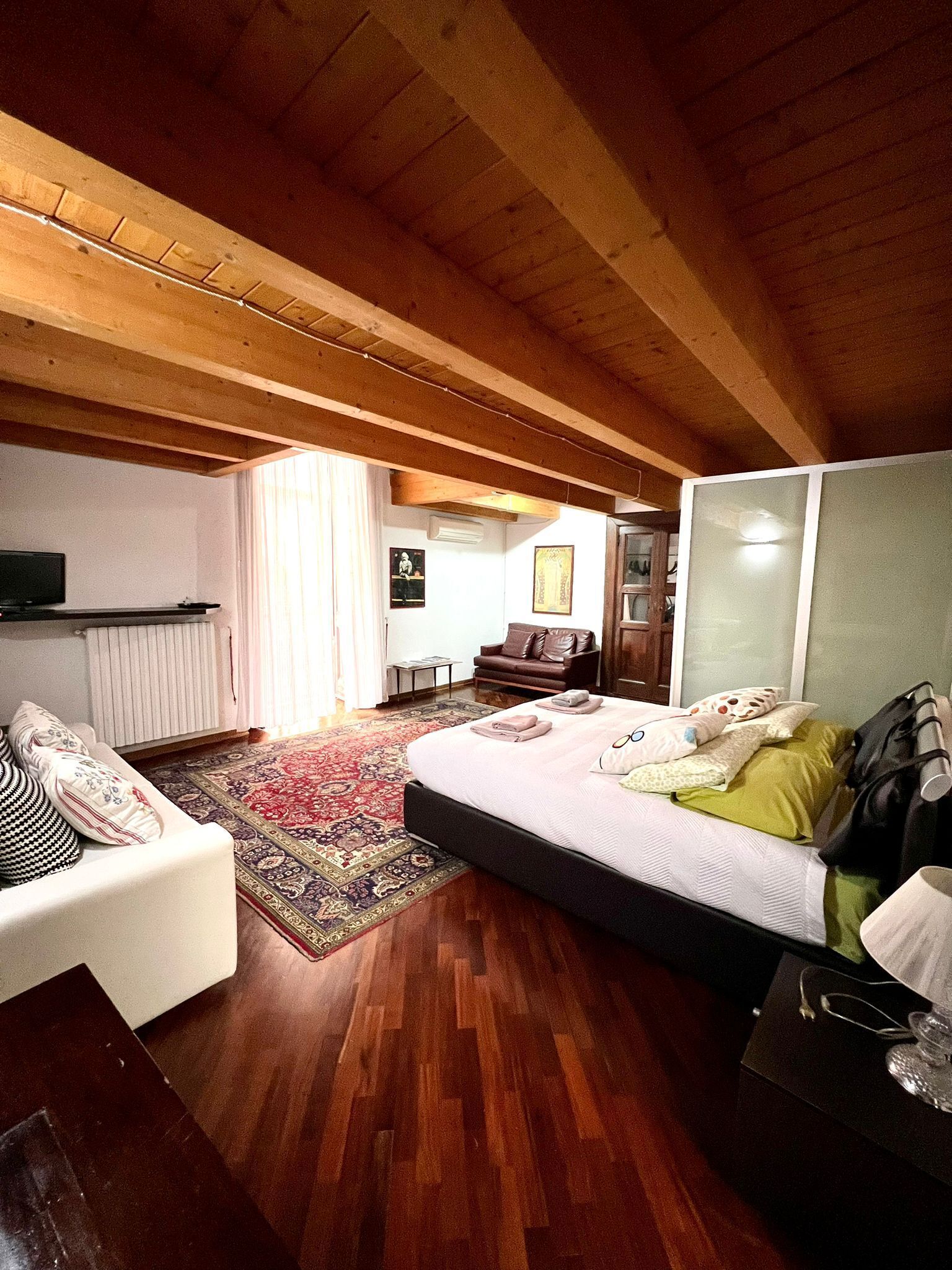 A bedroom with a wooden ceiling, a bed with pillows and a blanket, a sofa with cushions, a small table, and a window with curtains.