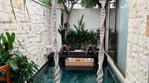 Una pequeña piscina al aire libre con una silla mecedora de madera, rodeada de muros de piedra y abundante vegetación.