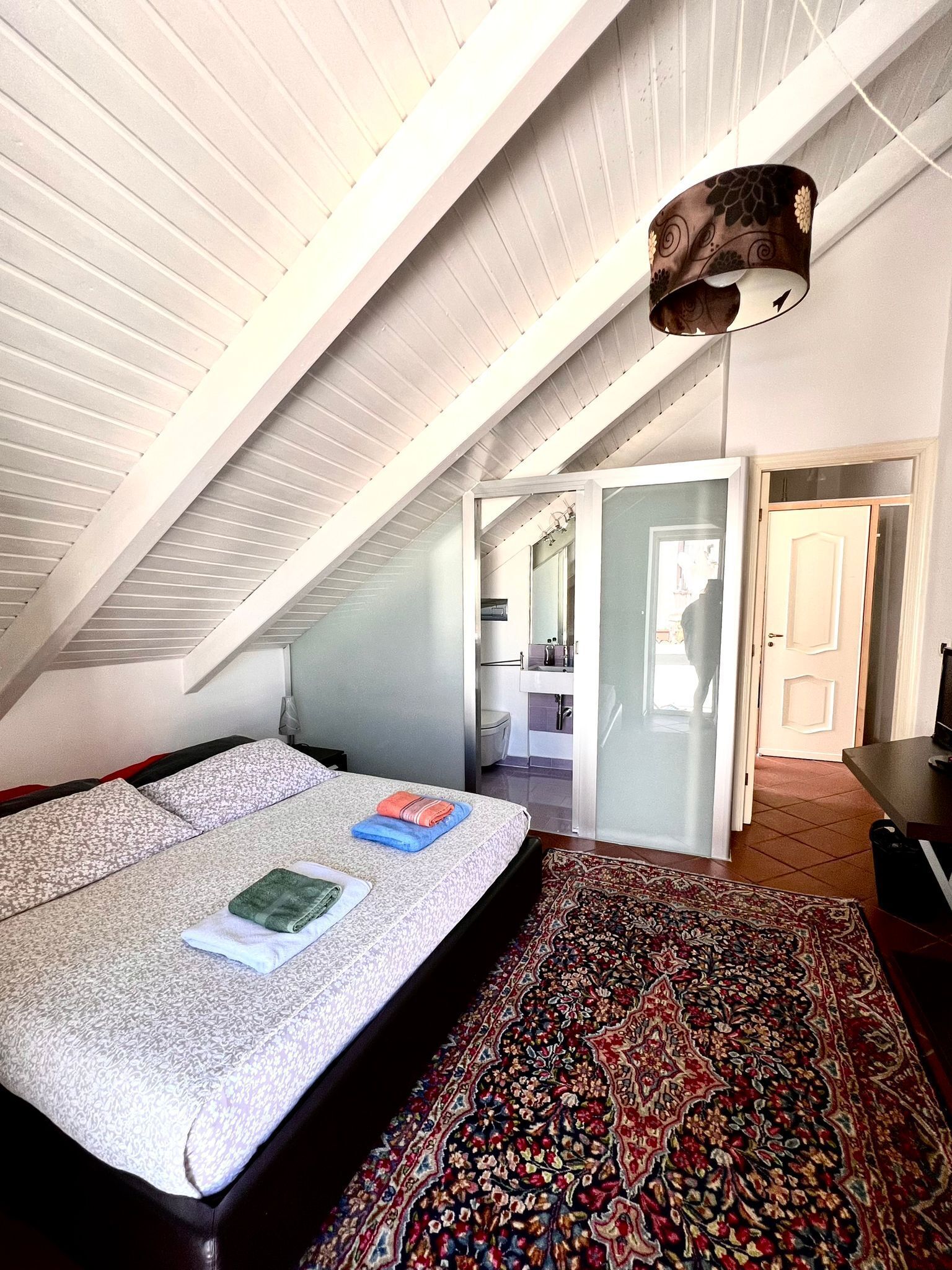 A bedroom with a sloped ceiling, a bed with a patterned bedspread, a bedside table, and a large, ornate rug.