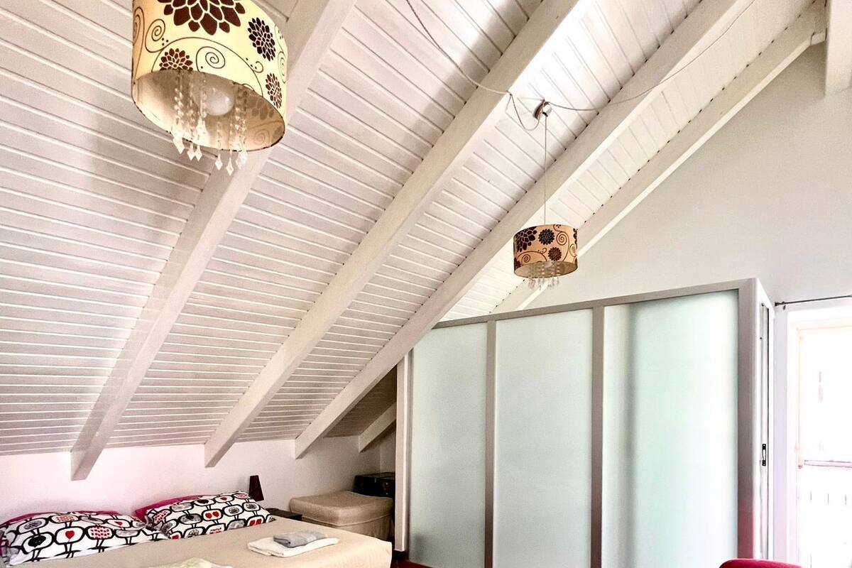 Un dormitorio con una cama, una silla roja, un tapete estampado y un techo con vigas a la vista.