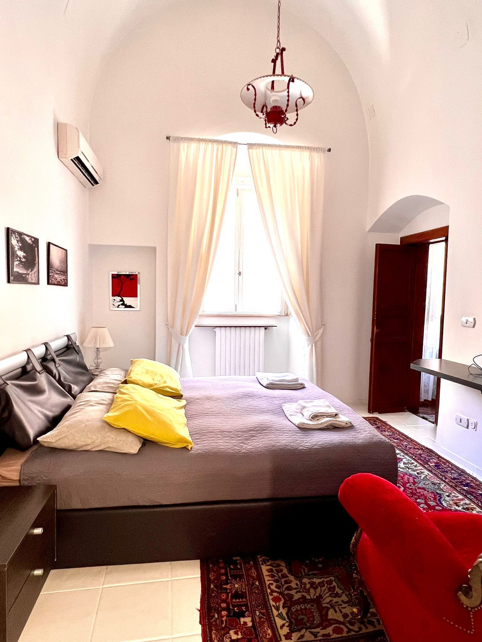 A bedroom with a bed, a red chair, a hanging light fixture, and a window with curtains.