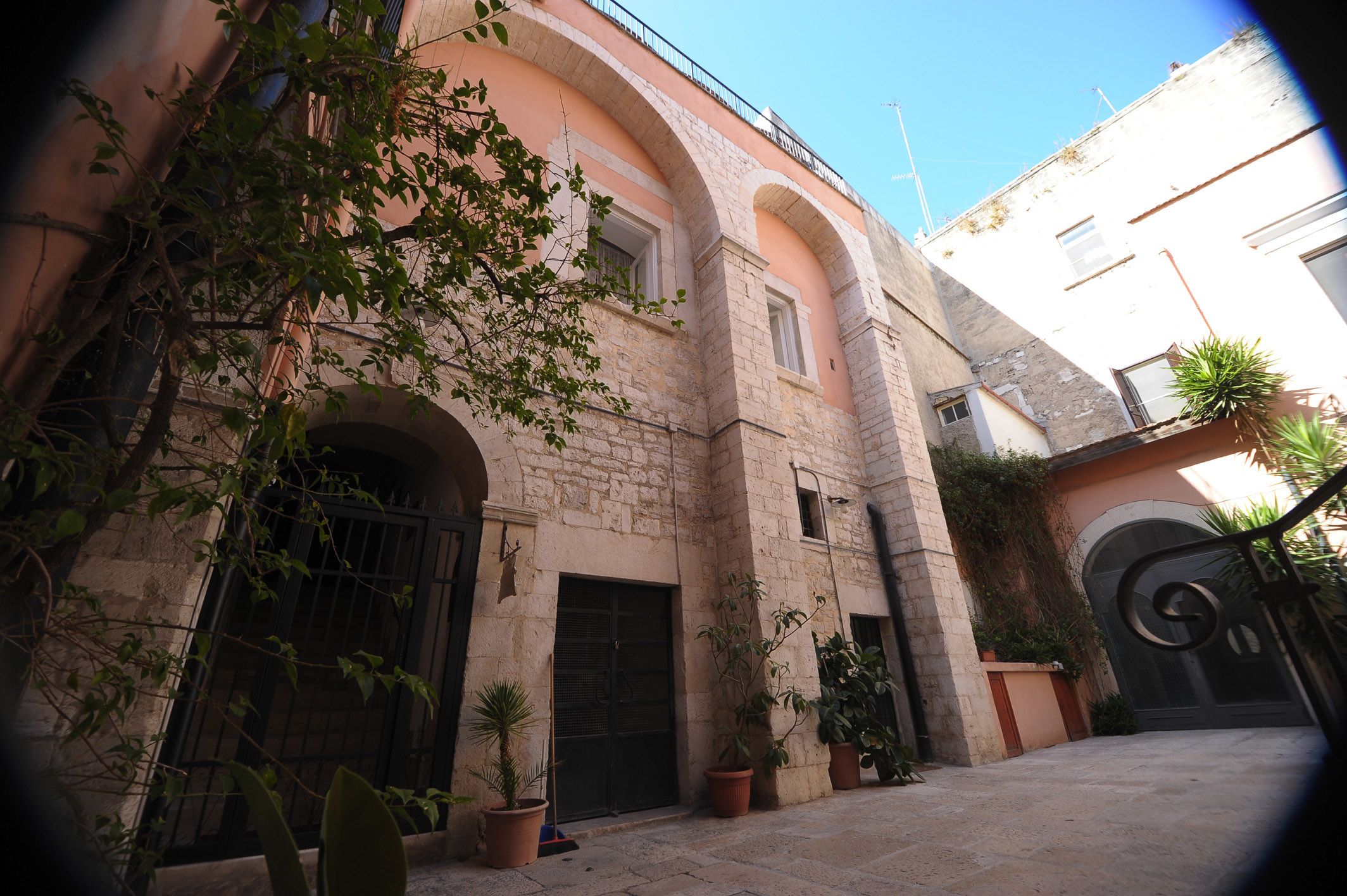 A courtyard with a stone building featuring arched doorways and windows, surrounded by potted plants and a metal gate.
