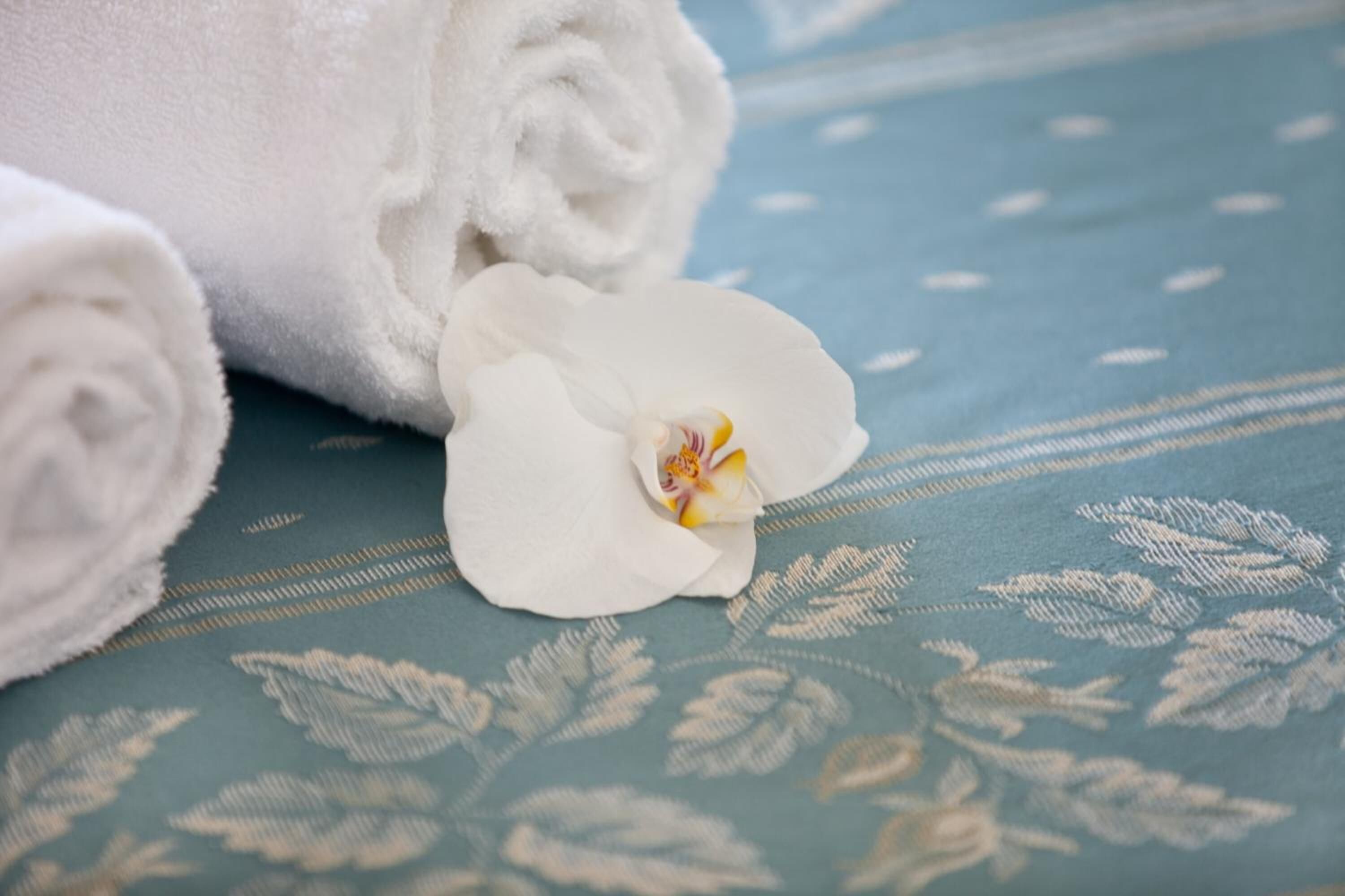 Une serviette blanche roulée et une orchidée blanche posées sur un tissu bleu à motifs.