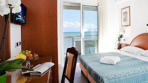 Habitación de hotel con cama, mesita de noche, escritorio con planta, silla, vista al océano y televisor en la pared.
