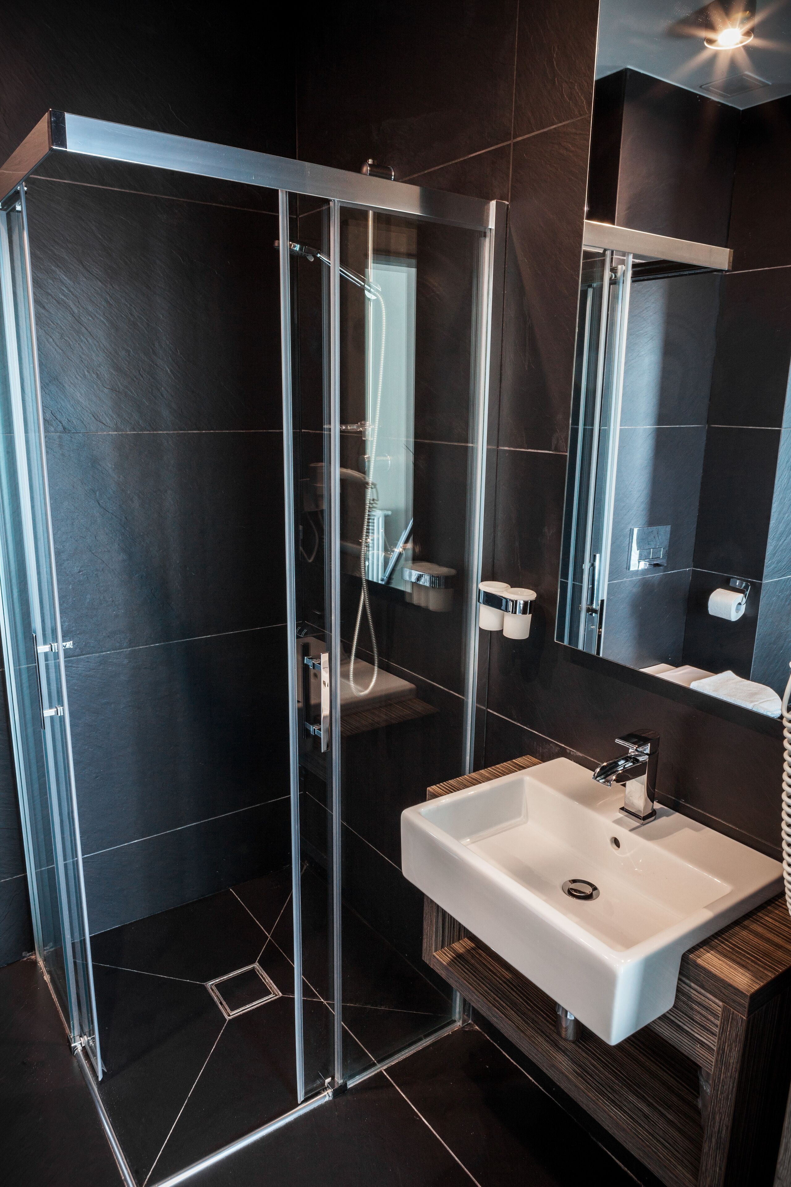 A modern bathroom with a glass-enclosed shower, a white sink, and dark tiled walls.