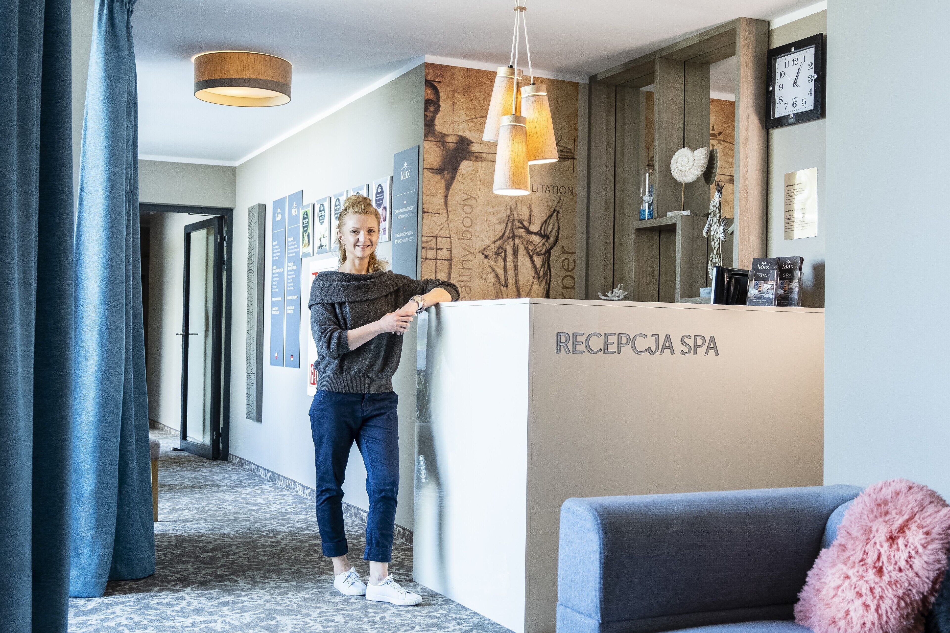 Ein Empfangsbereich mit einer Rezeption, die mit „RECEPTION SPA“ beschriftet ist, einer Wand mit gerahmten Bildern, einer Uhr und einem Sofa mit einem flauschigen Kissen.