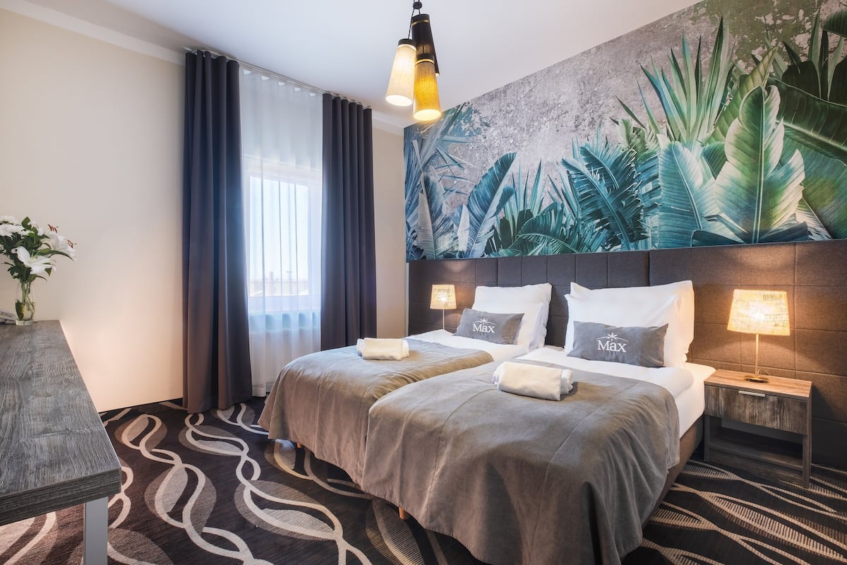 A hotel room with two beds, a large tropical mural, a modern chandelier, and a view of the city through the window.