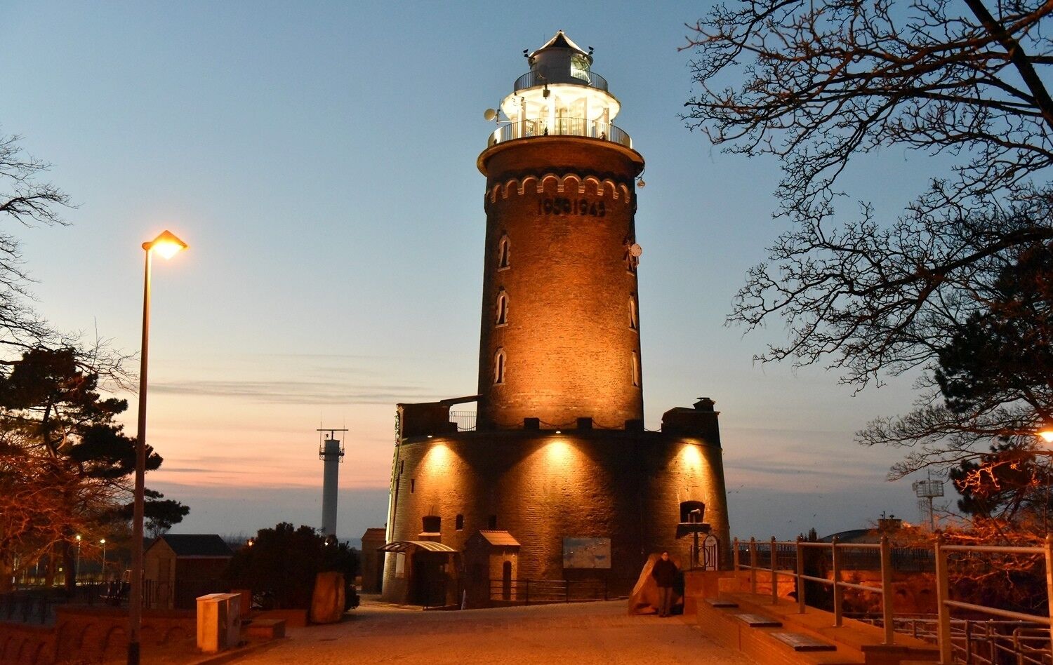 A brick lighthouse with a light on top, surrounded by trees and a paved walkway.