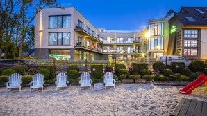 A modern building with multiple balconies and a sandy area with Adirondack chairs. - Max Health Resort SPA (Ustronie Morskie)