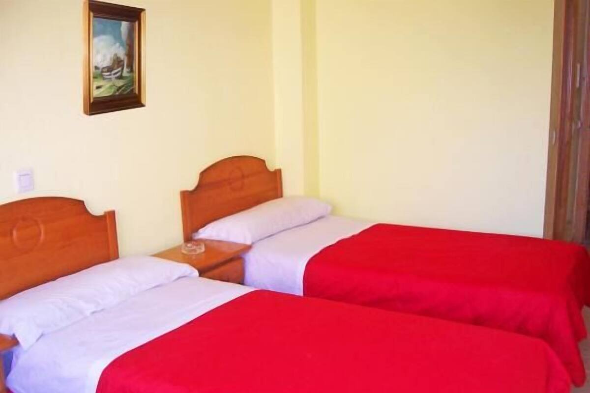 Una habitación con dos camas de madera, cada una con una colcha roja y almohadas blancas. Hay una mesita de noche con una lámpara entre las camas.