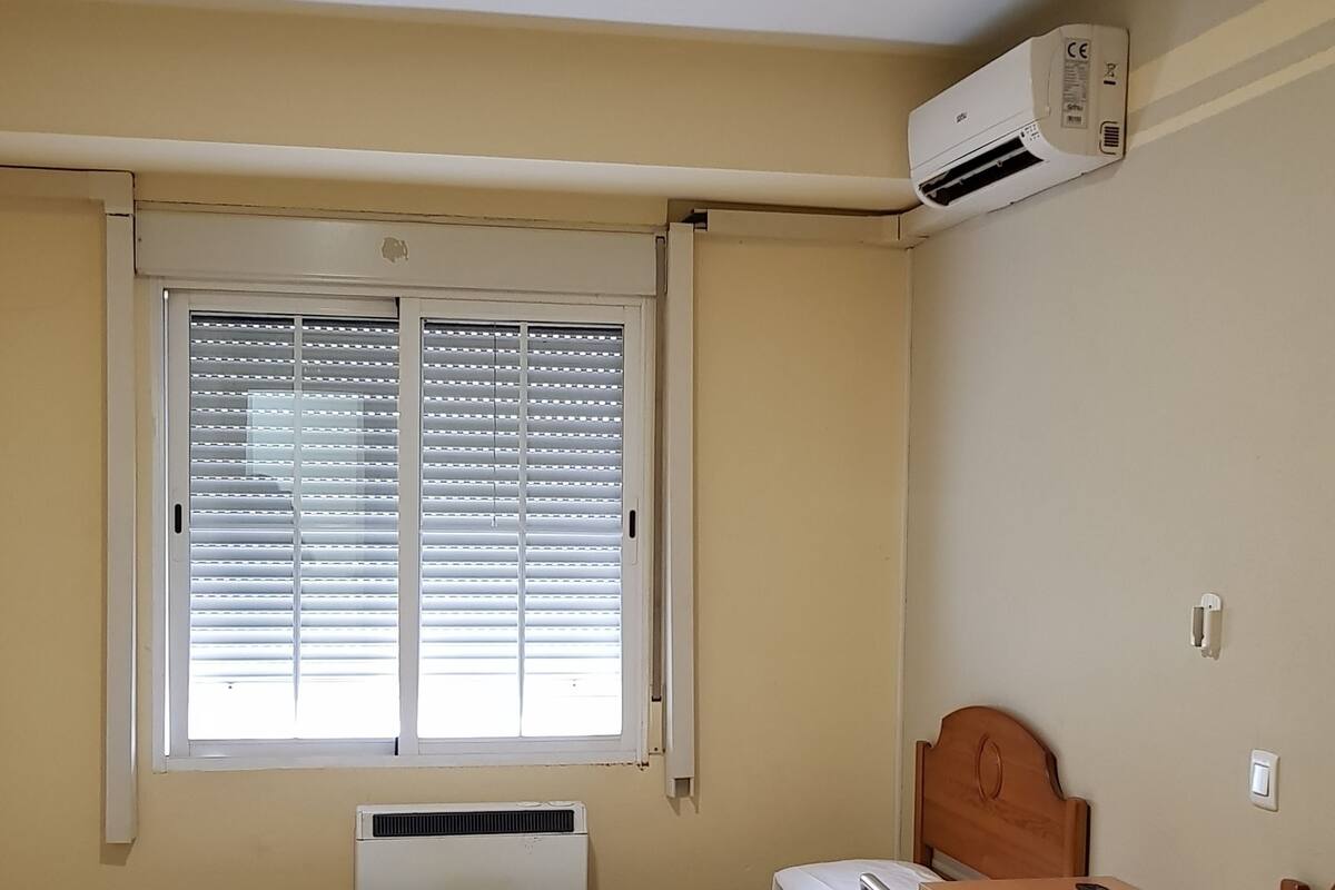 Una habitación con dos camas, una ventana con persianas, un equipo de aire acondicionado y un escritorio con silla.