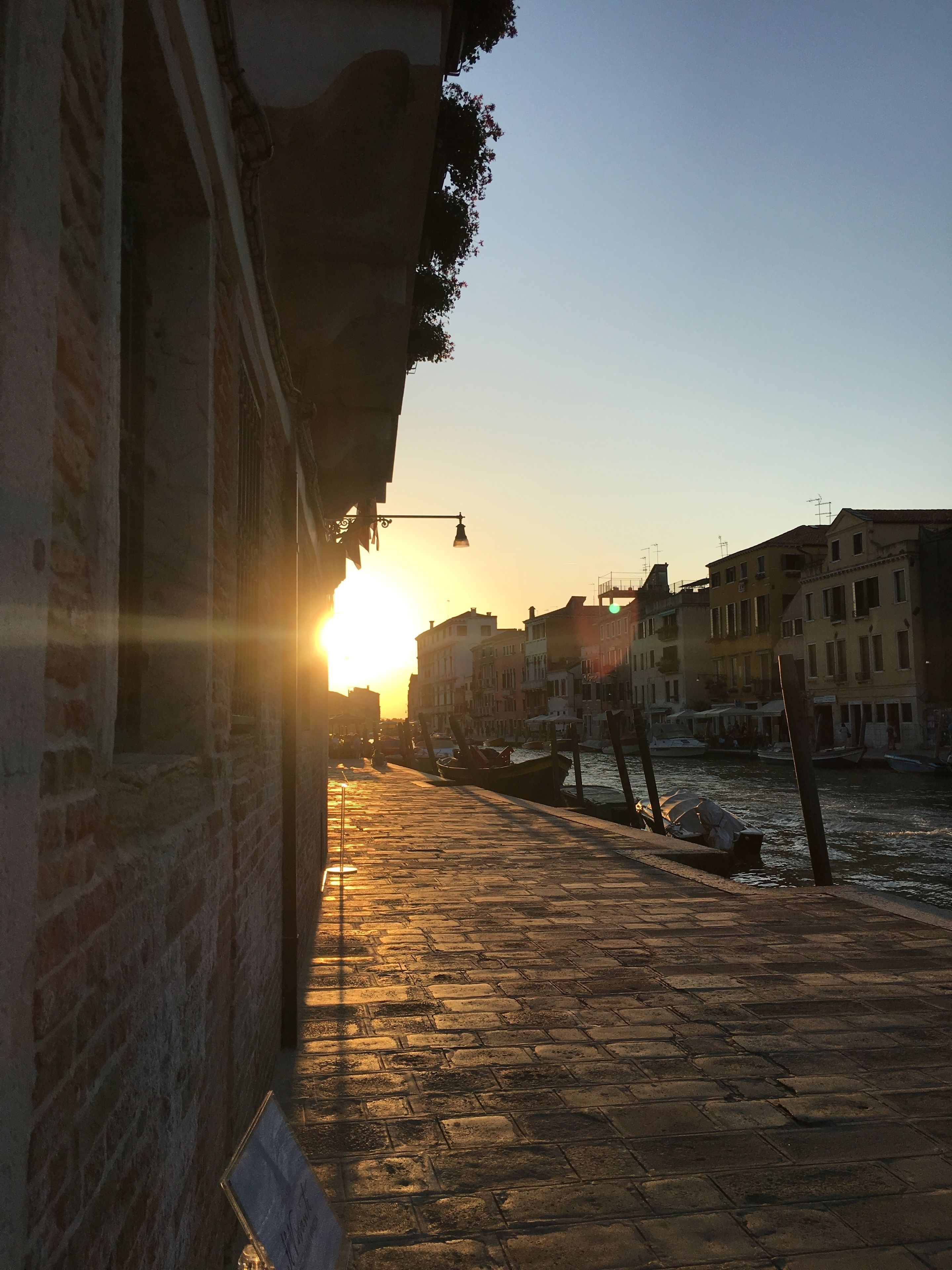 A sunset view of a canal with boats, buildings, and a cobblestone walkway.