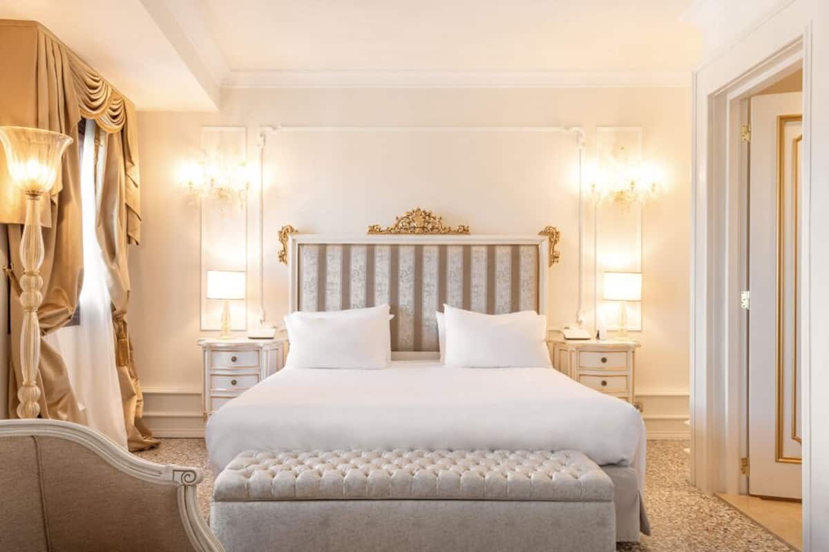 A large, elegantly furnished bedroom with a tufted headboard, a large bed, two bedside tables with lamps, and a large mirror.