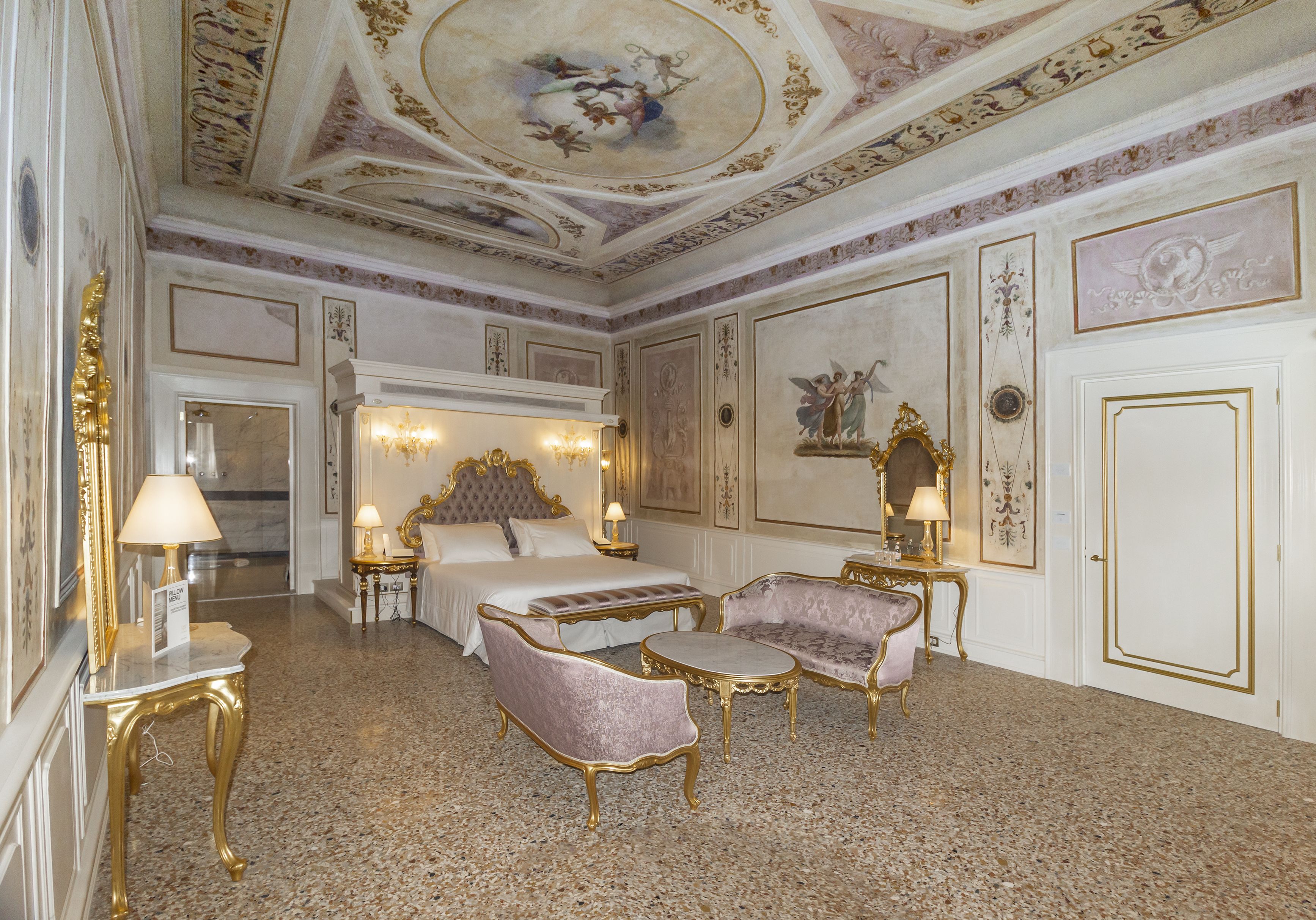 Une chambre d’hôtel luxueuse, dotée d’un grand lit, de deux fauteuils, d’une petite table et d’un plafond orné de motifs artistiques complexes.
