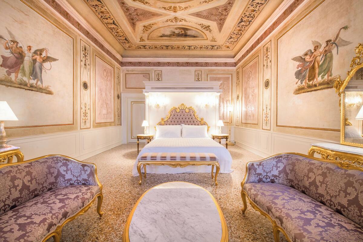 A luxurious bedroom with a large bed, ornate ceiling, and classical wall paintings.