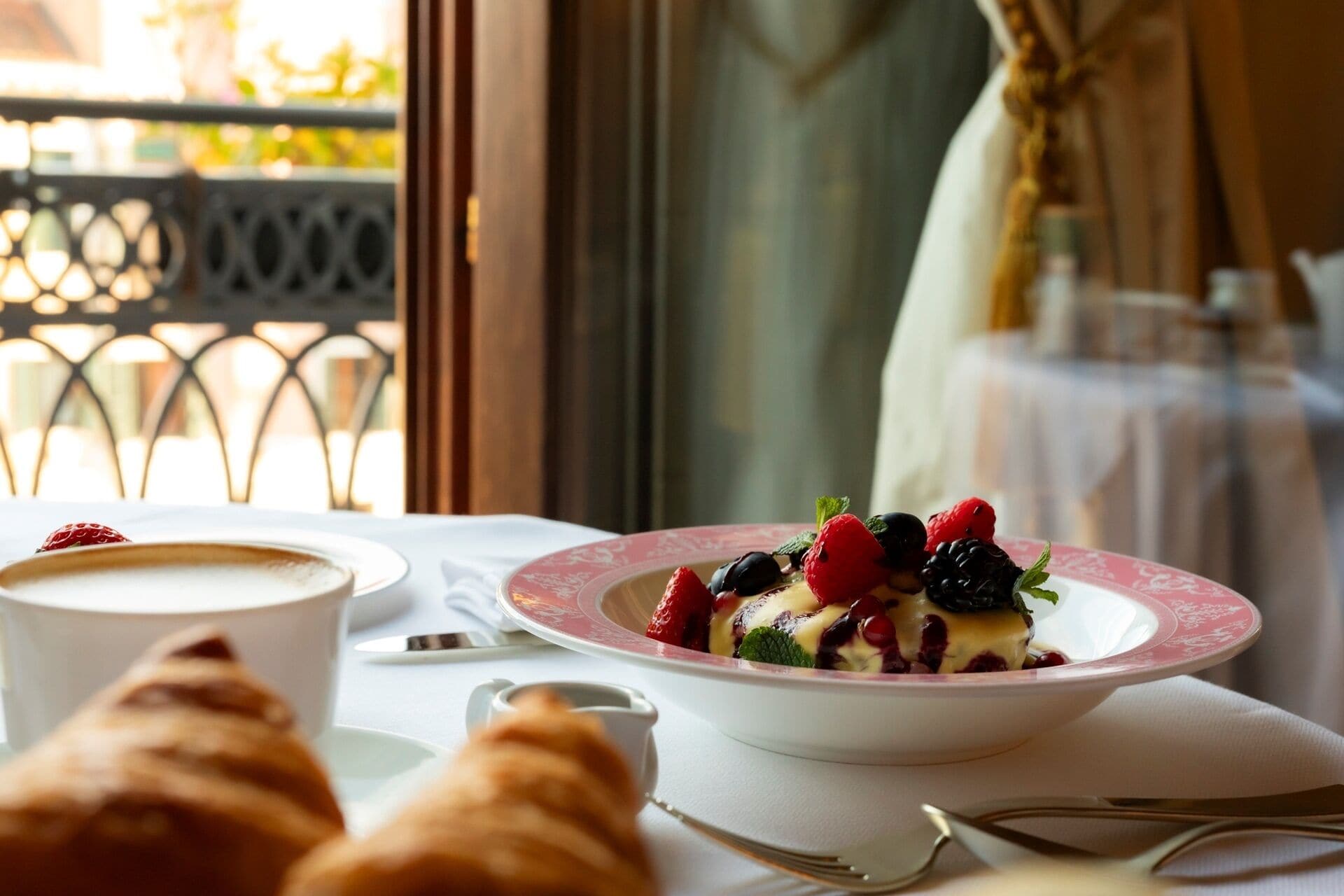 Une table à manger avec une assiette à dessert garnie de fruits rouges et un croissant, une tasse de café et un petit verre d’eau.