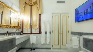 Et moderne badeværelse med marmor bordplader, et stort spejl, et badekar og et fjernsyn der viser et bybillede.