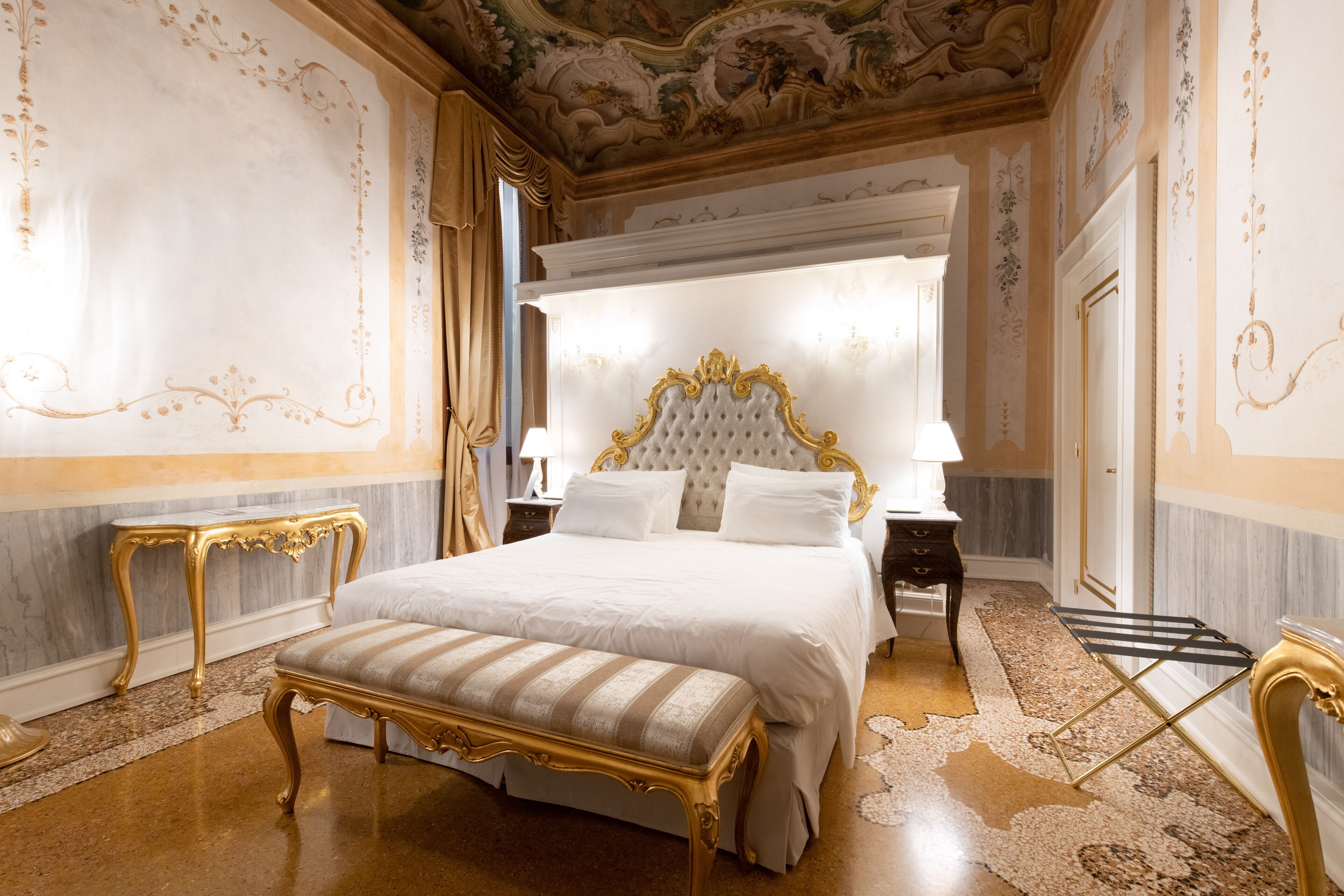 Une chambre à coucher luxueuse, dotée d’un grand lit, de meubles dorés richement décorés et d’une fresque murale au plafond aux détails soignés.