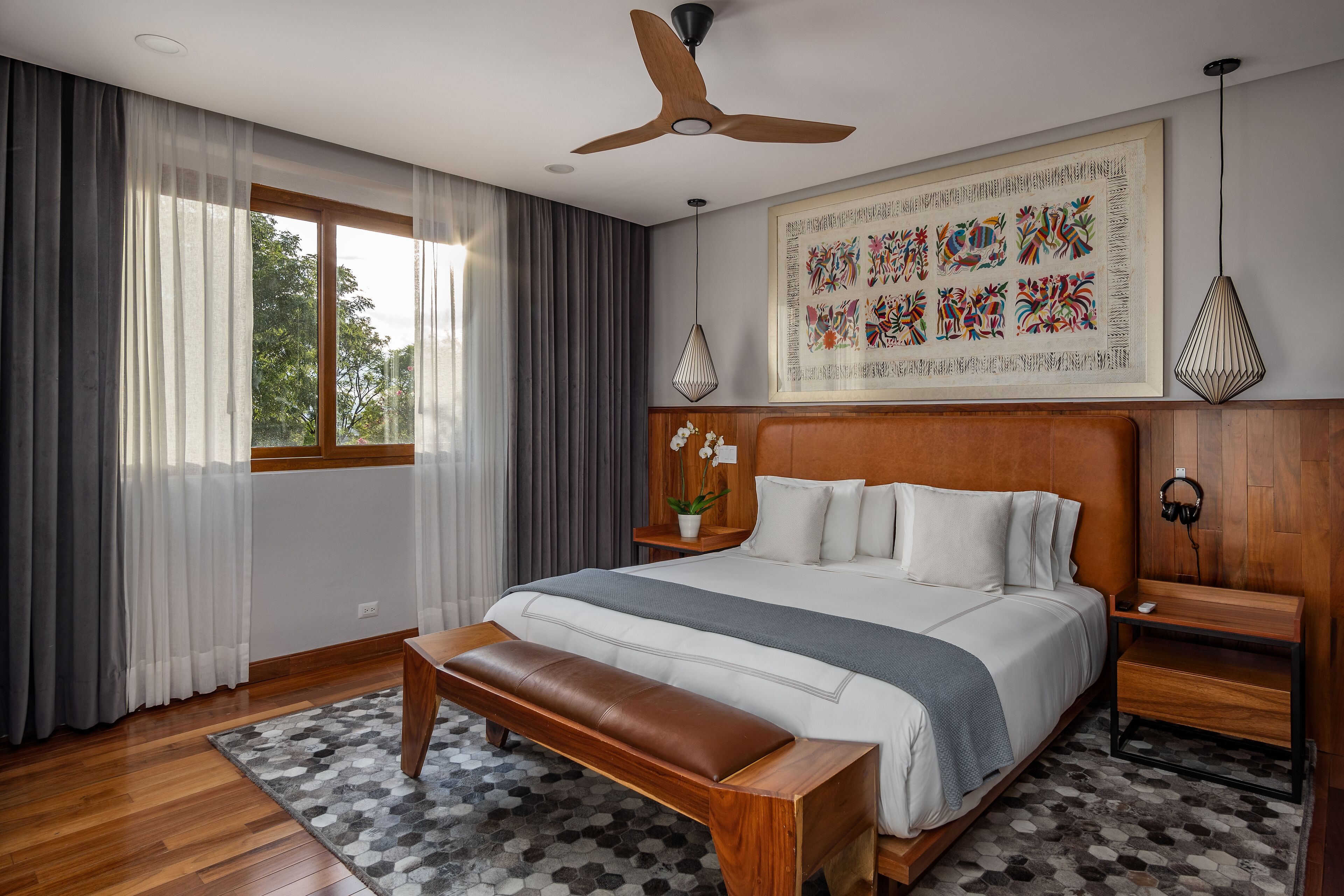A modern bedroom with a large bed, a wooden bench, a ceiling fan, a window with curtains, a wall-mounted artwork, and a nightstand with a lamp.