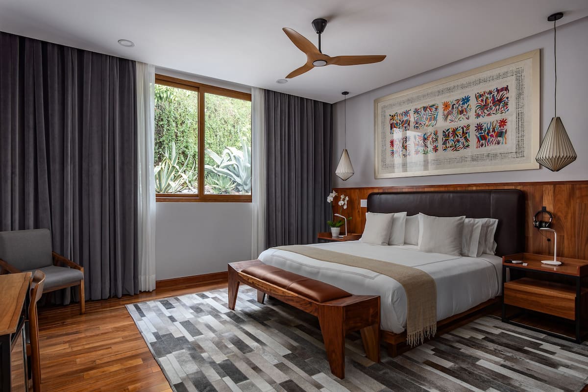 A modern bedroom with a large bed, a wooden bench, a desk, and a window with curtains.
