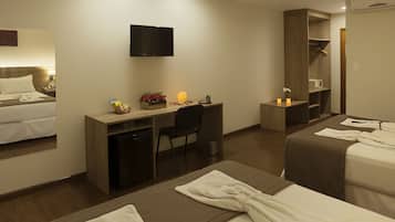 Quarto de hotel com cama, escrivaninha, cadeira, televisão, guarda-roupa e pequena mesa com vela e flores.
