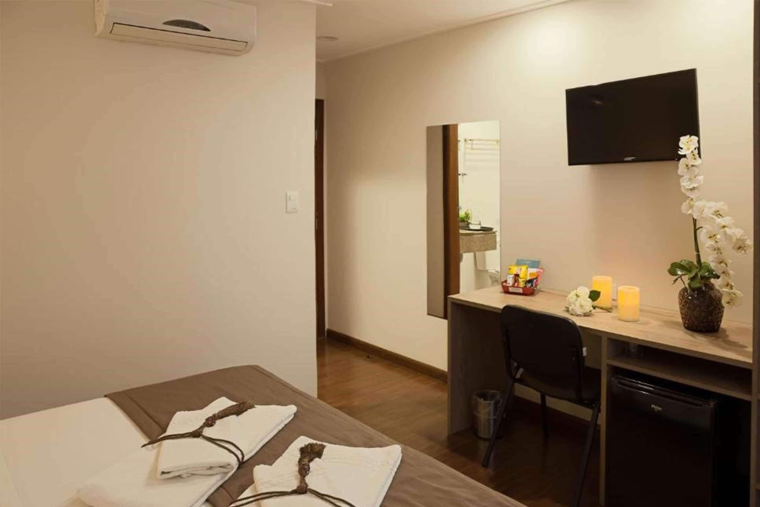 Habitación de hotel con cama, escritorio, silla, televisor y una mesita con velas y flores.