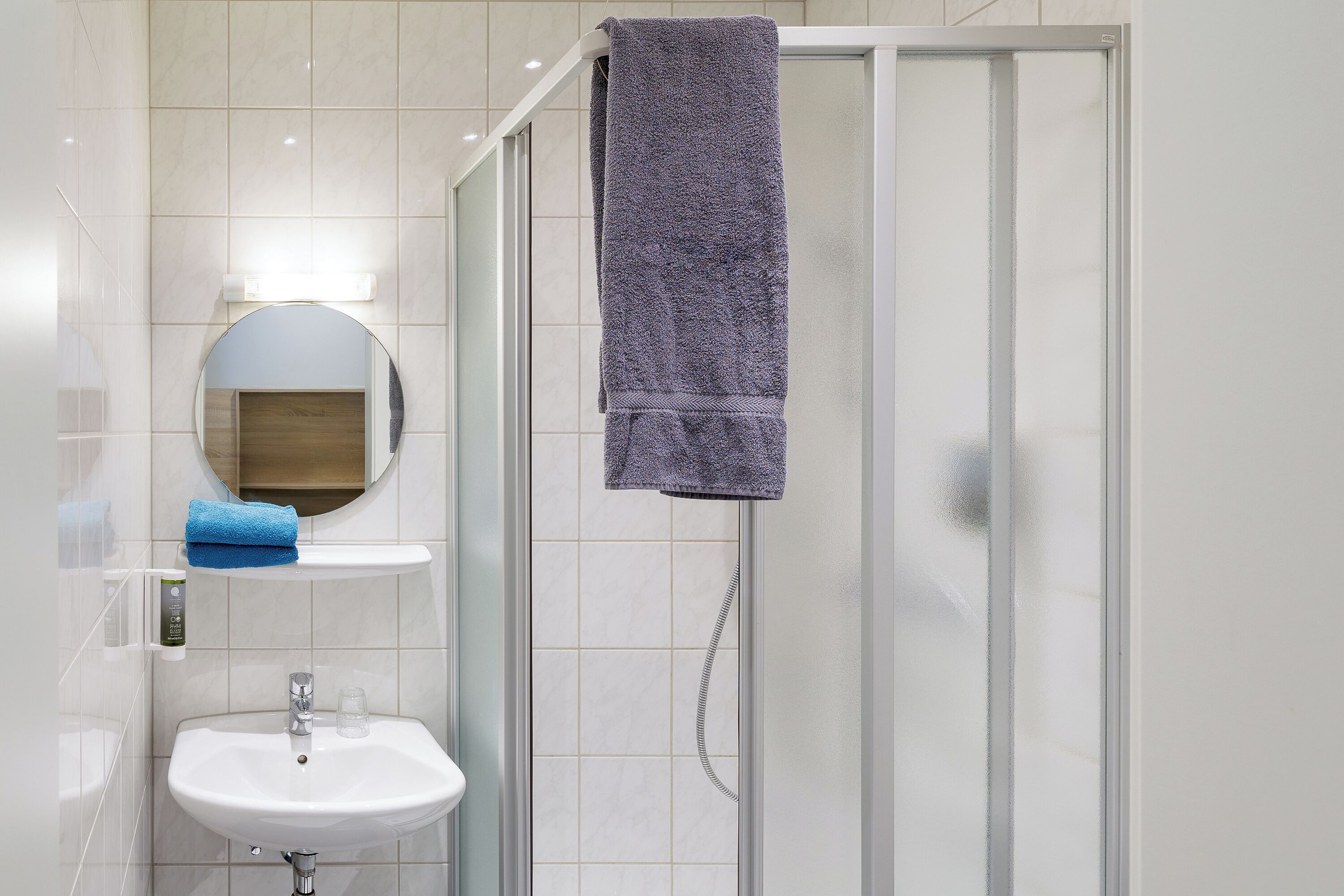 A bathroom with a glass shower enclosure, a sink, and a mirror with a towel hanging on the wall.