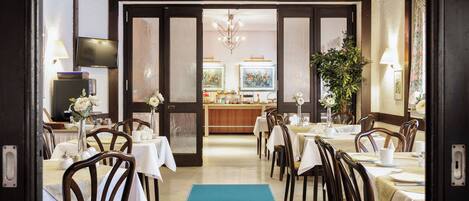 Comedor de hotel con mesas dispuestas para los huéspedes, un bar al fondo y una planta grande en el centro.