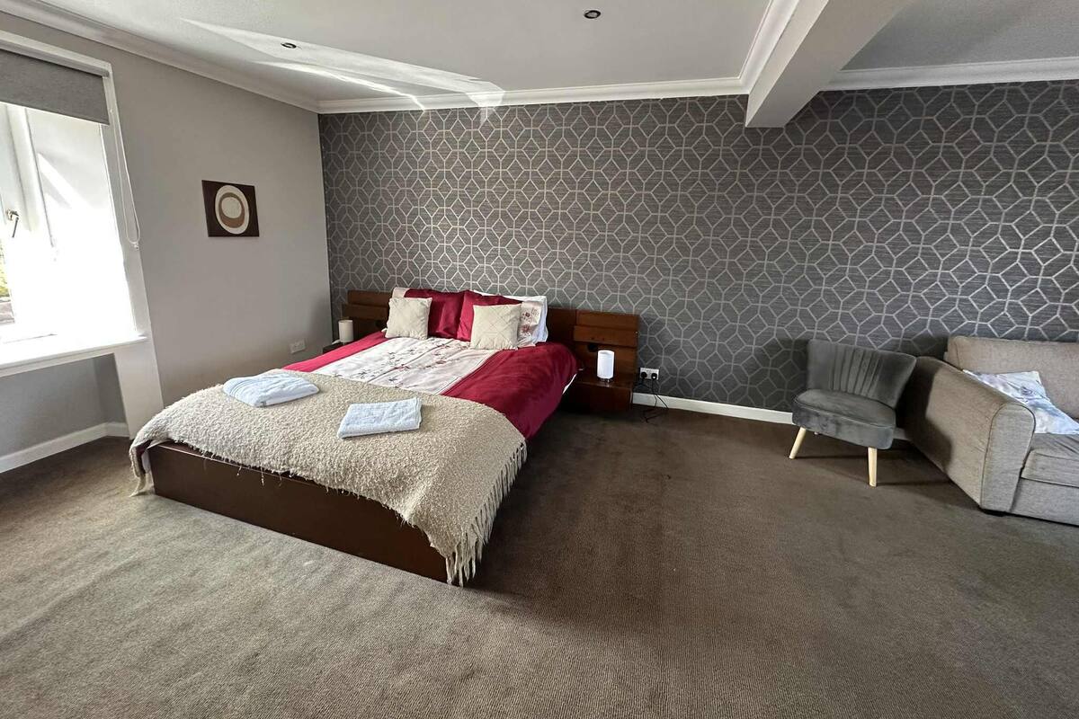 A hotel room with a large bed, a grey patterned wall, a grey armchair, and a window with curtains.