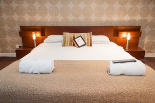 Cama bem arrumada com travesseiro, duas luminárias de cabeceira e duas toalhas enroladas sobre um tapete texturizado.