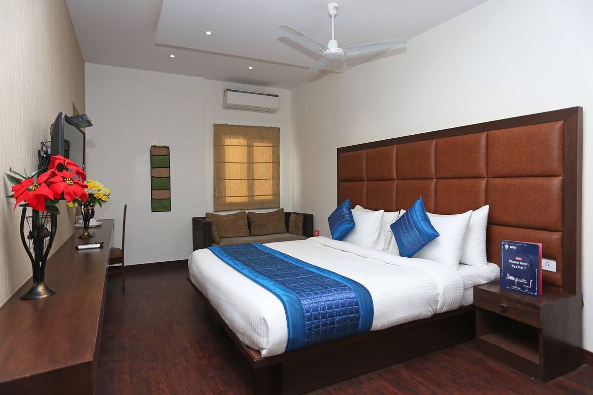 A hotel room with a large bed, a bedside table, a nightstand, a desk with a chair, a mirror, and a window with blinds.