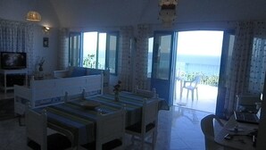 A room with a dining table, chairs, a TV, and a view of the sea through large windows. - Dar Tomoko & Salah (Raf Raf)