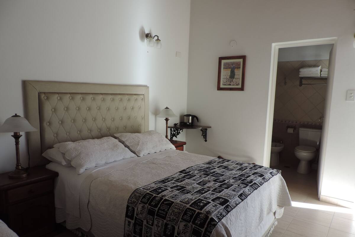 Un lit bien fait, recouvert d’une couverture à motifs, une lampe de chevet, un tableau encadré au mur et une vue sur la salle de bain.