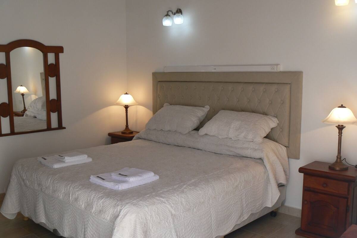 Un lit bien fait, avec une tête de lit, deux lampes de chevet, un miroir et une petite table de nuit en bois avec une lampe.