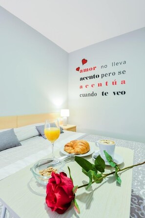 Una habitación de hotel con una cama, una mesa de desayuno con cruasanes, un vaso de jugo de naranja, una rosa roja y una pared con una cita de amor.