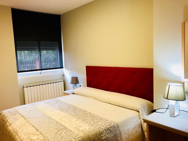 Un dormitorio con cabecera roja, una cama con ropa de cama estampada, una mesita de noche con lámpara y una ventana con persianas.