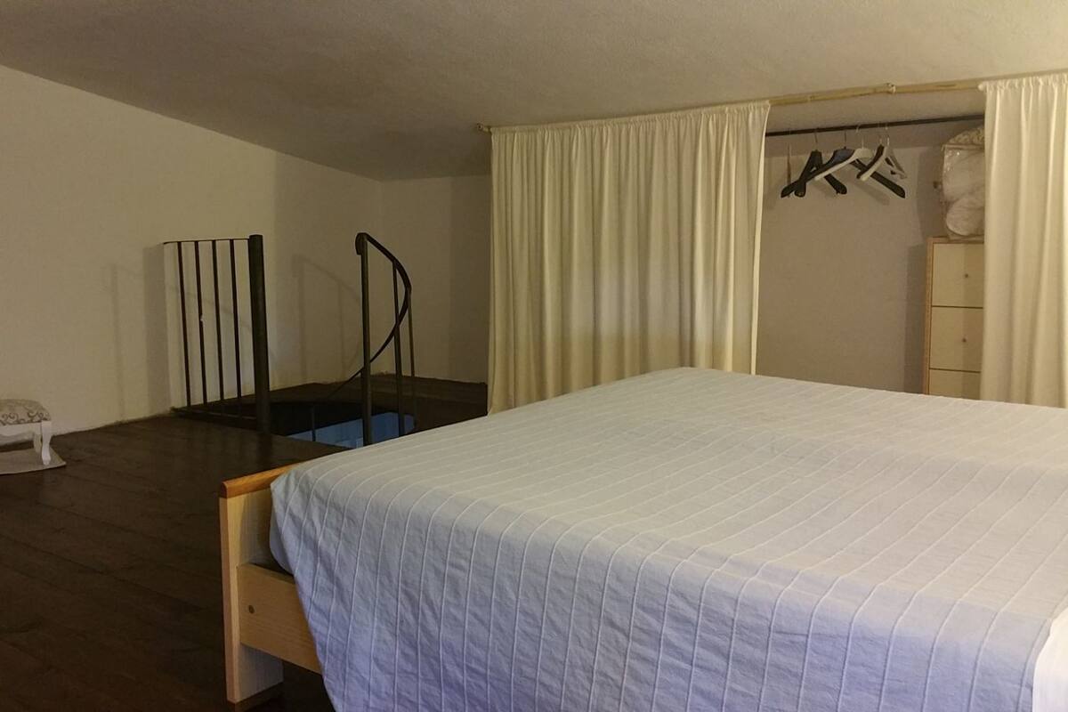 A bedroom with a bed, a small bench, a chair, a wardrobe, and a window with curtains.