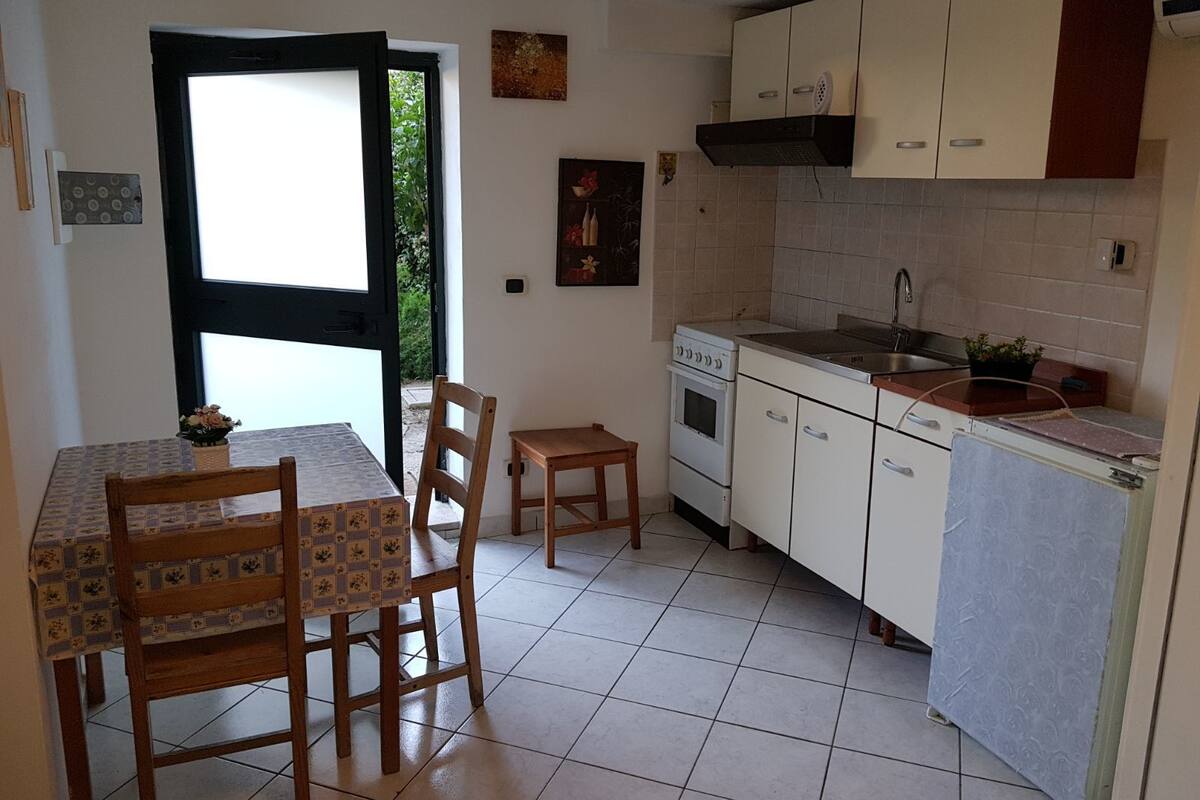 A compact kitchen with white cabinets, a stove, and a sink. There is a dining table with chairs and a window with a view of greenery.