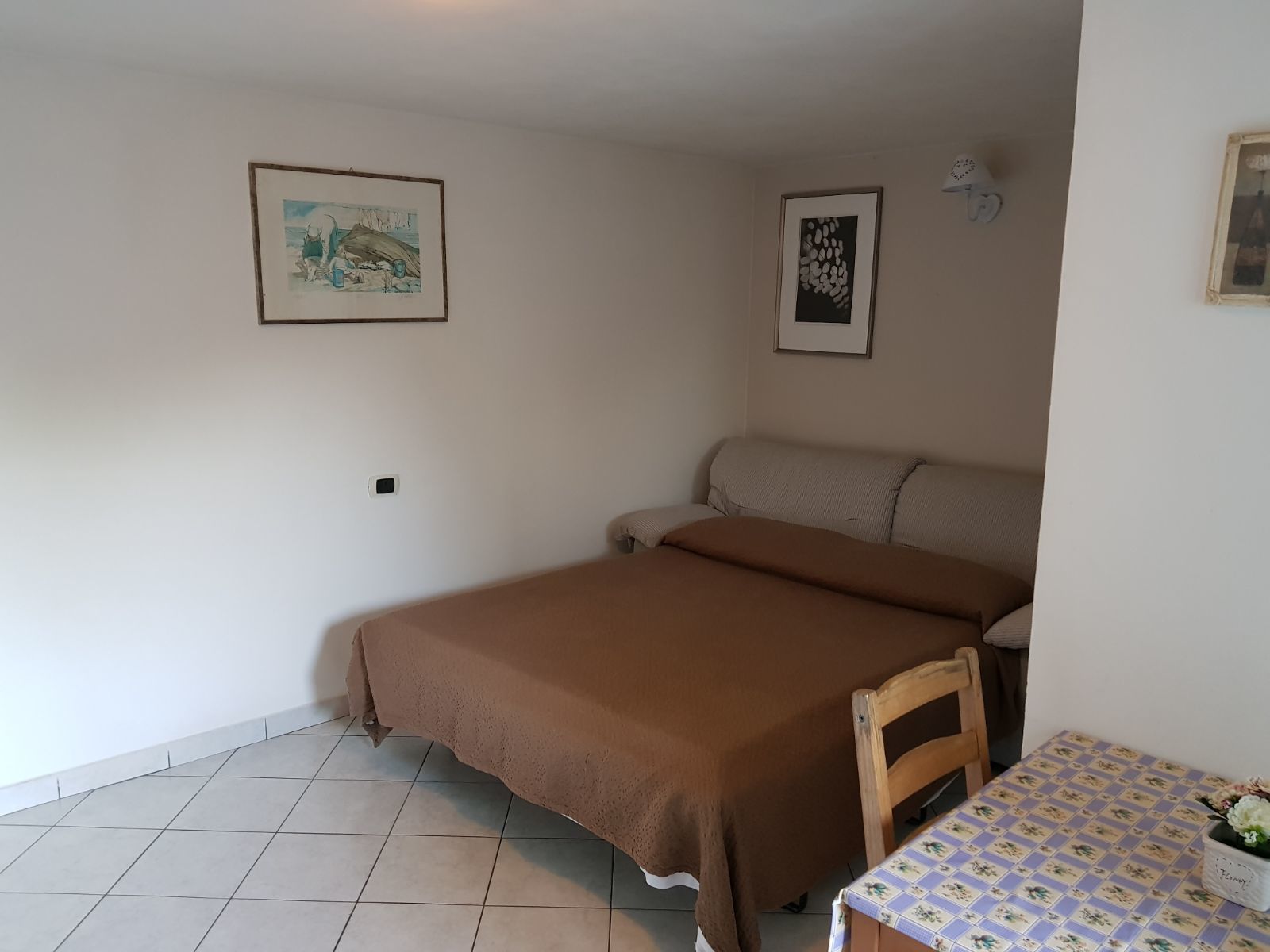 A bedroom with a bed, a small table with a floral tablecloth, and two framed pictures on the wall.