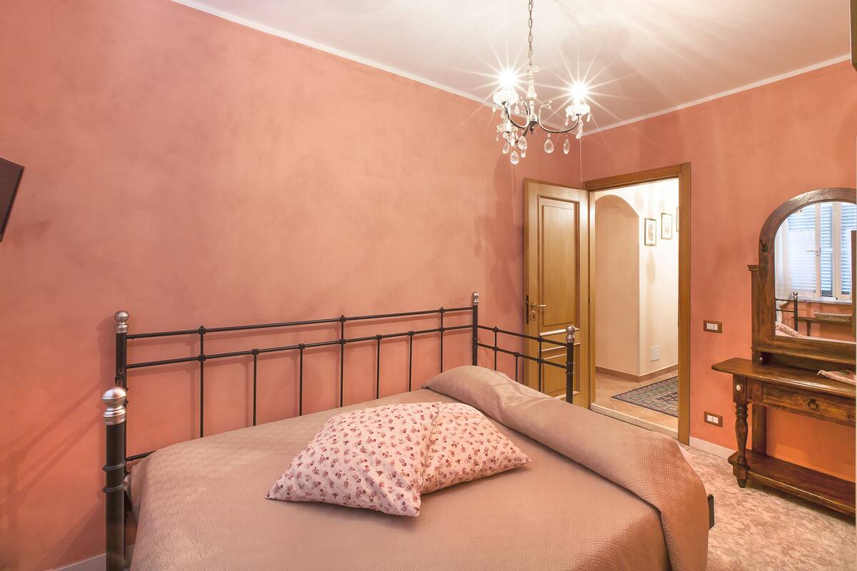 A bedroom with a bed, a chandelier, a wooden dresser, and a door leading to another room.